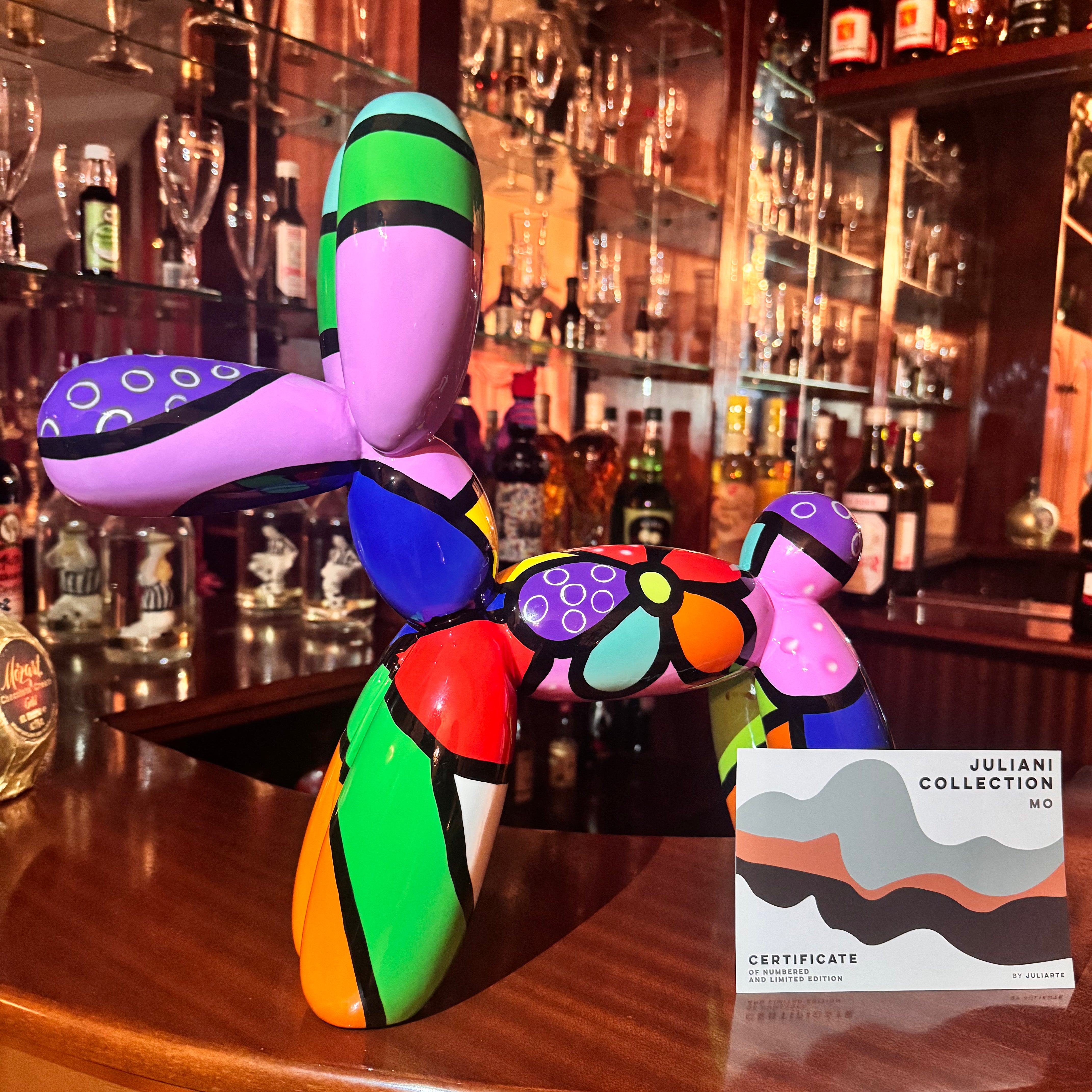 A colorful Pop Art balloon dog sculpture displayed on a wooden bar counter in a bar setting, next to a certificate card from the Juliani Collection.