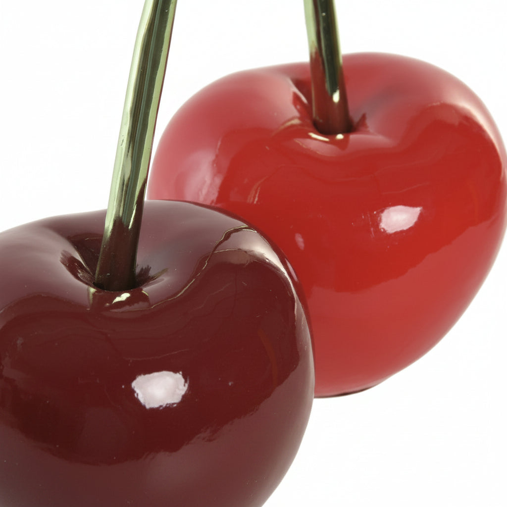 Two glossy cherry-shaped decorative objects with dark green stems, one deep red and one dark burgundy, shown close up