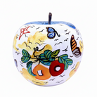 A decorative ceramic apple figurine with a white base, hand-painted with colorful motifs including green leaves, red and orange fruits, blue butterflies, black birds, and yellow clouds, featuring a dark brown stem.