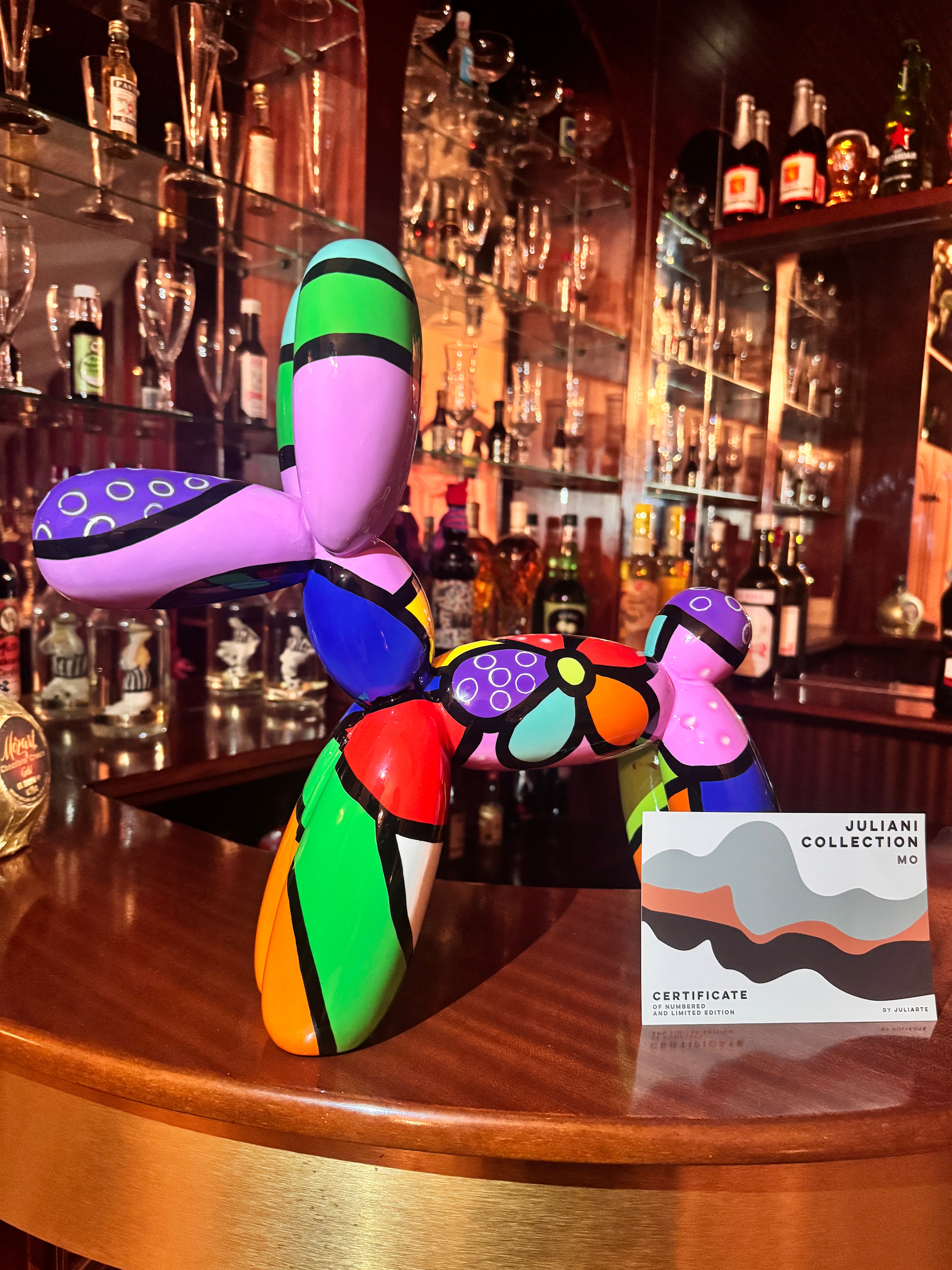 A colorful Pop Art balloon dog sculpture displayed on a wooden bar counter in a bar setting, next to a certificate card from the Juliani Collection.