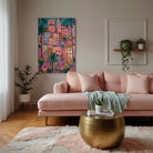 A colorful framed wall art print depicting a tropical hillside village with pink buildings and palm trees, displayed above a blush pink sofa in a modern living room setting.